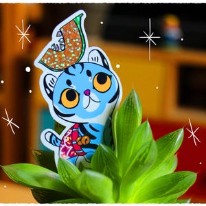 May include: A blue and white cartoon tiger wearing a green and orange watermelon hat. The tiger is standing in a green succulent plant.