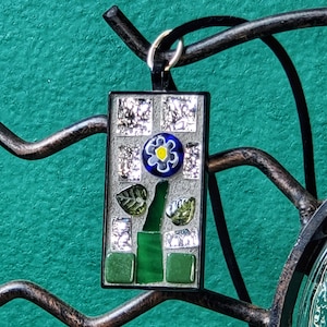 May include: A rectangular pendant featuring a mosaic design with a central blue flower with a yellow center, green glass stem and leaves, and silver-toned reflective tiles set against a light gray background, framed in a thin black border.