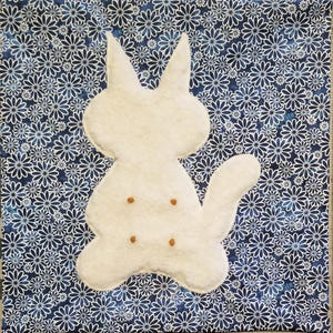 May include: A square fabric panel featuring a dark blue background with a repeating white daisy pattern. Centered on the panel is a white, plush, cat-shaped applique with two pointed ears, a curved tail, and four small, brown, circular accents.