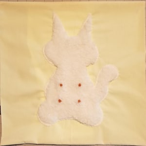 May include: A square light yellow fabric cushion cover featuring a centered white plush cat silhouette appliqué. The cat shape has four small, evenly spaced orange circular accents on its body, creating a simple and clean visual design.