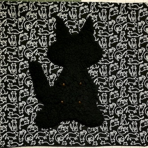 May include: A black cat-shaped pillow with four gold-colored buttons, set against a black and white cat-themed fabric background. The fabric features a repeating pattern of white cat illustrations on a black backdrop.