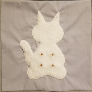 May include: A light gray square fabric with a white cat applique. The cat is made of a textured material and has four small brown dots. The cat has pointed ears and a curved tail. The fabric is a solid color.