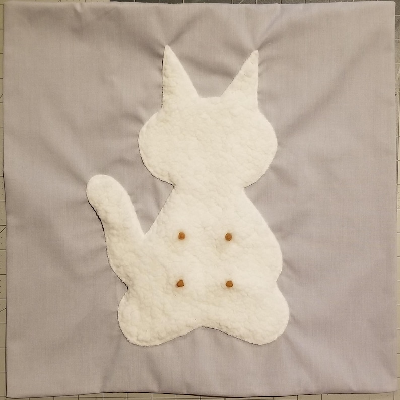 May include: A light gray square fabric with a white cat applique. The cat is made of a textured material and has four small brown dots. The cat has pointed ears and a curved tail. The fabric is a solid color.