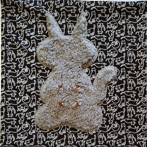 May include: A gray, textured, plush cat-shaped pillow featuring four small, round, orange-colored buttons on its front. The pillow is positioned against a black fabric background that is covered in a repeating white line-art pattern of cats.