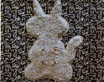 May include: A gray, textured, plush cat-shaped pillow featuring four small, round, orange-colored buttons on its front. The pillow is positioned against a black fabric background that is covered in a repeating white line-art pattern of cats.