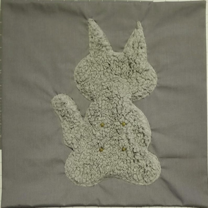May include: A gray square pillow with a plush, light gray cat applique. The cat has button eyes and is made of a fuzzy material. The pillow is a solid gray color.
