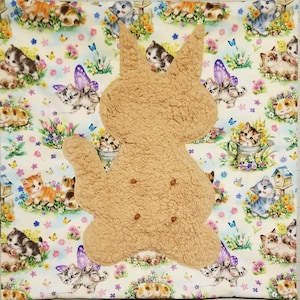 May include: A tan, textured, sherpa-like fabric cutout in the shape of a cat silhouette with four small brown circular accents, layered over a light-colored fabric background featuring a repeating pattern of kittens, butterflies, and colorful flowers.