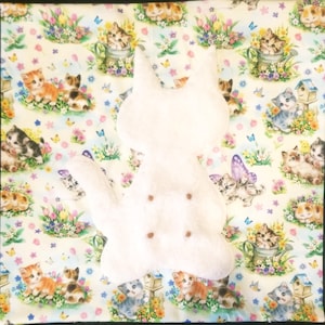 May include: A white cat-shaped plush toy rests on a patterned fabric. The fabric features a repeating design of kittens, flowers, butterflies, and birdhouses in a pastel color palette. The plush toy has a soft, textured appearance.