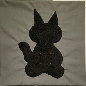 May include: A gray square pillow with a black cat silhouette. The cat is made of a textured material and has three small, gold-colored buttons. The pillow is a solid gray color.