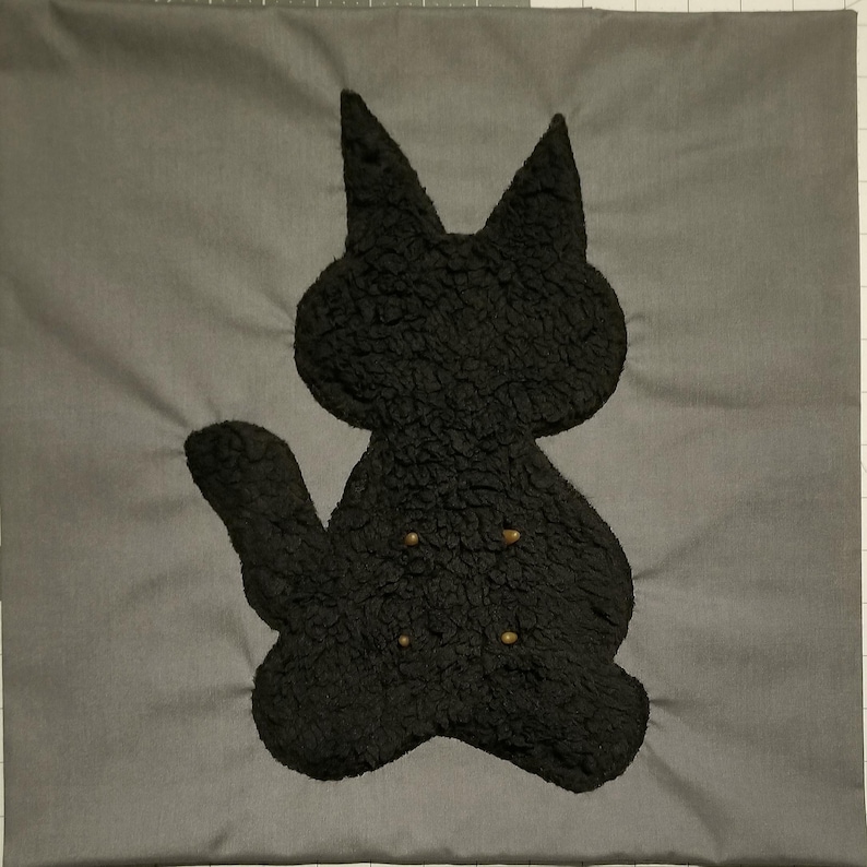 May include: A gray square pillow with a black cat silhouette. The cat is made of a textured material and has three small, gold-colored buttons. The pillow is a solid gray color.