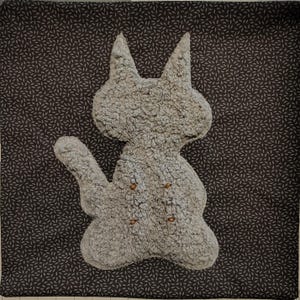 May include: A gray plush bunny-shaped pillow with button details. The pillow is on a dark gray fabric with a repeating leaf pattern. The bunny has long ears and an outstretched arm.