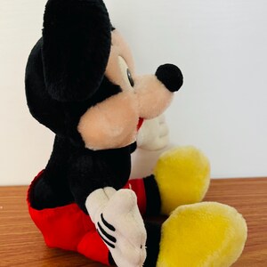 May include: A plush Mickey Mouse toy, featuring black fur, a tan face, and large black ears. The toy wears a red shorts, white gloves, and yellow shoes. The classic character is in a seated position.