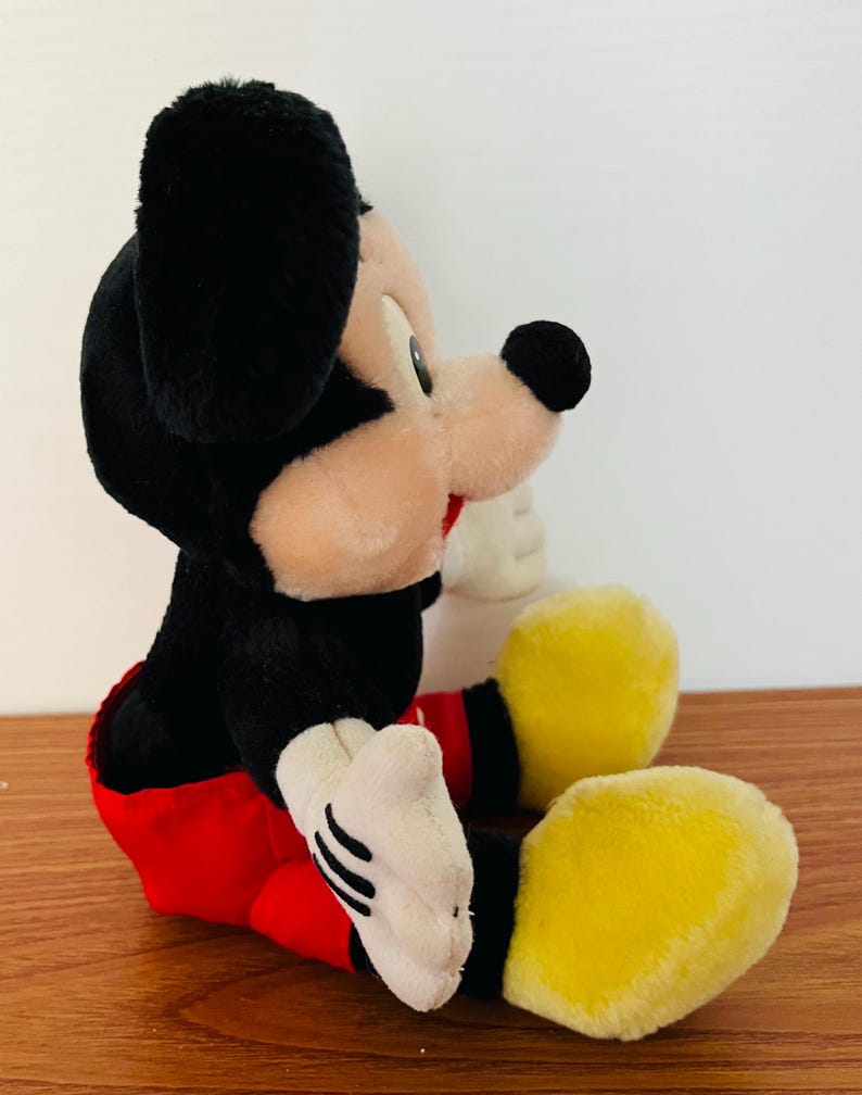 May include: A plush Mickey Mouse toy, featuring black fur, a tan face, and large black ears. The toy wears a red shorts, white gloves, and yellow shoes. The classic character is in a seated position.