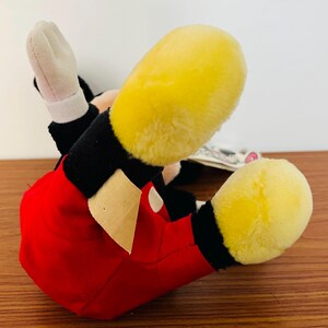 May include: A plush Mickey Mouse toy, lying on its back, with yellow feet, red shorts, black arms and white gloves. The toy has a tag attached. The background is a wooden surface.