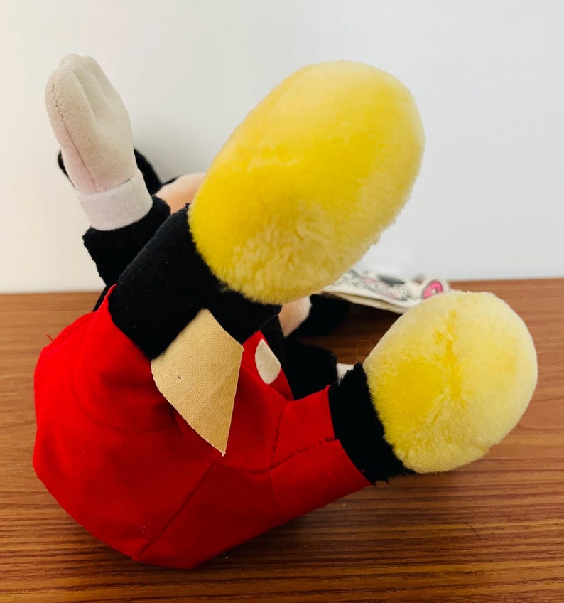 May include: A plush Mickey Mouse toy, lying on its back, with yellow feet, red shorts, black arms and white gloves. The toy has a tag attached. The background is a wooden surface.