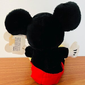 May include: A plush Mickey Mouse toy, viewed from the back, with large black ears, a black body, red shorts, and white gloves. A tag is attached with a caution message. The toy is on a wooden surface.