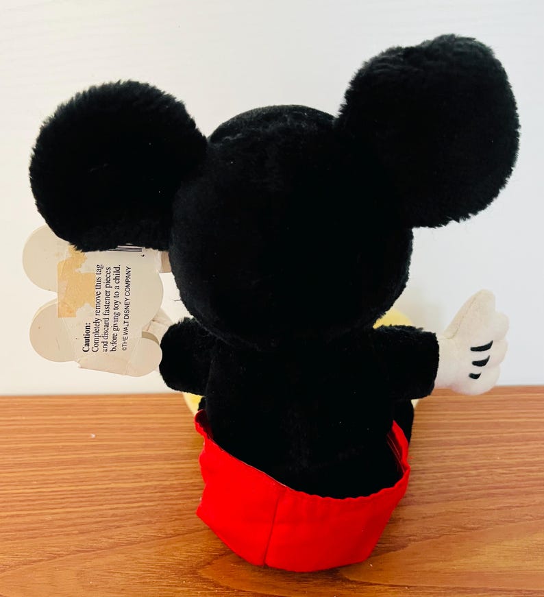 May include: A plush Mickey Mouse toy, viewed from the back, with large black ears, a black body, red shorts, and white gloves. A tag is attached with a caution message. The toy is on a wooden surface.