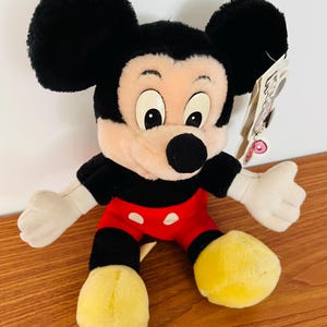 May include: A plush Mickey Mouse toy with black ears, a black body, red shorts, and yellow shoes. The toy has a tag attached. The toy is a classic Disney character.