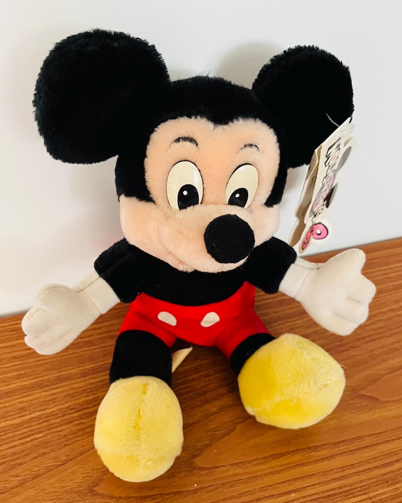 May include: A plush Mickey Mouse toy with black ears, a black body, red shorts, and yellow shoes. The toy has a tag attached. The toy is a classic Disney character.