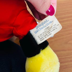 May include: A plush toy featuring a red body, black accents, and a yellow foot. The tag reads "Disneyland Walt Disney World" and includes the Disney logo and product information.