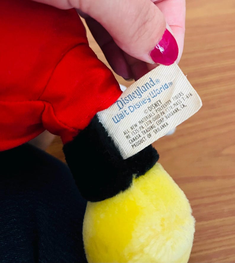May include: A plush toy featuring a red body, black accents, and a yellow foot. The tag reads "Disneyland Walt Disney World" and includes the Disney logo and product information.