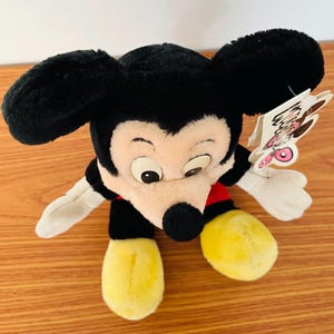 May include: A plush Mickey Mouse toy with black ears, a black nose, and a cream-colored face. The toy has yellow feet, white gloves, and a red and black outfit. A tag is attached to the toy.