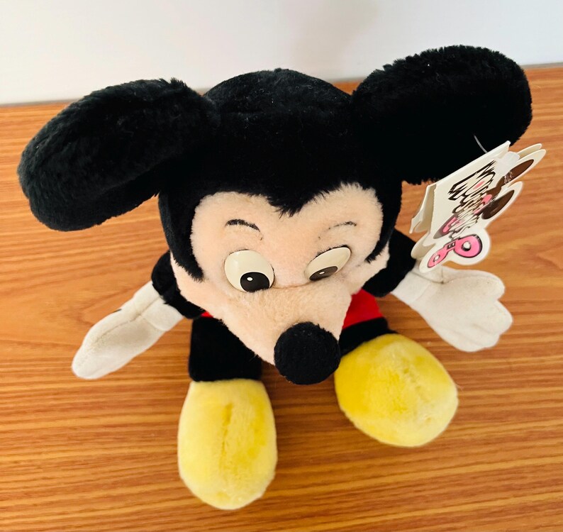May include: A plush Mickey Mouse toy with black ears, a black nose, and a cream-colored face. The toy has yellow feet, white gloves, and a red and black outfit. A tag is attached to the toy.