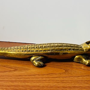 May include: A brass alligator figurine with a textured body and open mouth. The sculpture is detailed with visible teeth and legs, and is resting on a wooden surface.