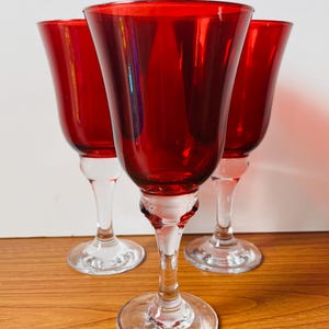 Puede incluir: Tres copas de vino rojas con tallos y bases transparentes dispuestas sobre una superficie de madera. Las copas tienen forma de tulipán y un acabado brillante. Los tallos son delgados y las bases circulares.