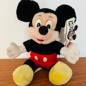 May include: A plush Mickey Mouse doll with black ears, a black shirt, red shorts with white buttons, and yellow feet. The doll has white gloves and a tag that reads "Mouseketeers".