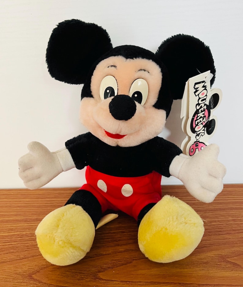 May include: A plush Mickey Mouse doll with black ears, a black shirt, red shorts with white buttons, and yellow feet. The doll has white gloves and a tag that reads "Mouseketeers".