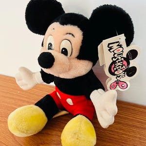 May include: A plush Mickey Mouse toy, featuring black ears, a black body, red shorts with white buttons, and yellow feet. The toy has a tag that reads "Mouseketeers".