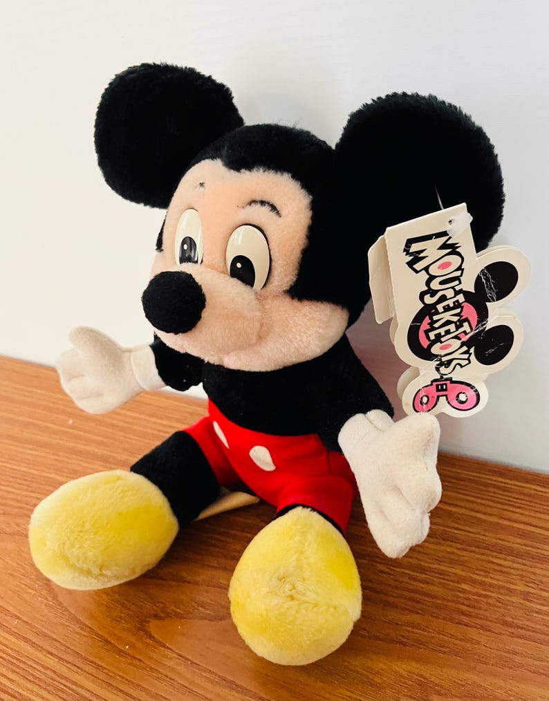 May include: A plush Mickey Mouse toy, featuring black ears, a black body, red shorts with white buttons, and yellow feet. The toy has a tag that reads "Mouseketeers".