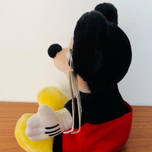 May include: A plush Mickey Mouse toy, featuring black fur, a red shorts, yellow shoes, and white gloves. The toy has a tag attached. The plush is sitting on a wooden surface.