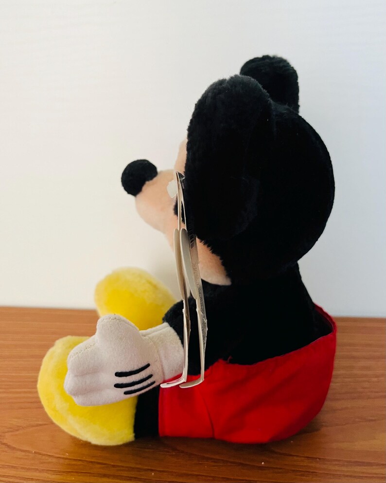 May include: A plush Mickey Mouse toy, featuring black fur, a red shorts, yellow shoes, and white gloves. The toy has a tag attached. The plush is sitting on a wooden surface.