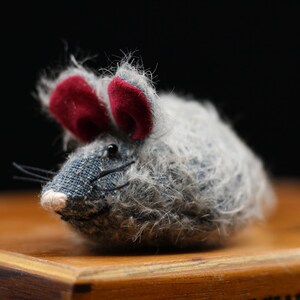 Puede incluir: Un juguete de ratón de peluche gris y blanco con orejas de fieltro rojo. El juguete está sentado sobre una superficie de madera.