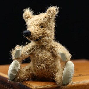 Pode incluir: Um pequeno urso de peluche castanho claro com patas brancas está sentado em uma caixa de madeira. A caixa tem o texto "25 Especiales Zechbauer Im" impresso nela.