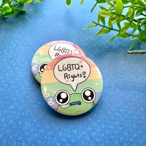 May include: Two round rainbow-colored buttons with a cartoon frog design. The top button says "LGBTQ+ Rights!" in a speech bubble. The bottom button says "LGBTQ" with a heart.