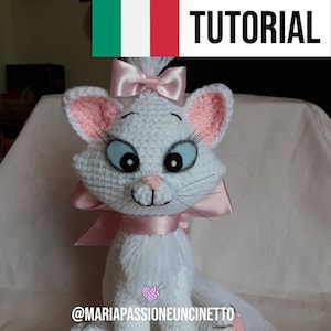 Può includere: Gatto bianco all'uncinetto con orecchie rosa, un fiocco rosa e occhi azzurri. Il gatto è seduto su una superficie bianca. Il testo "@mariapassioneuncinetto" è visibile nella parte inferiore dell'immagine.