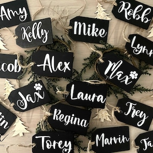 May include: Black chalkboard gift tags with white lettering, featuring names like Amy, Kelly, Mike, and Lynne. The tags are tied with twine and decorated with small wooden Christmas trees.