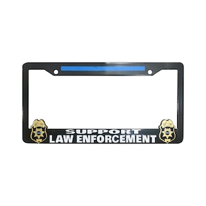May include: Black license plate frame with a blue stripe across the top. The words "SUPPORT LAW ENFORCEMENT" are printed in white. Gold-colored police badge emblems are on each side.
