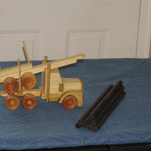 May include: A handcrafted wooden toy logging truck featuring a light tan finish, a flatbed trailer with vertical stakes, and five dark brown cylindrical wooden logs. The truck has six wheels, a cab, and a long boom arm extending from the rear section.