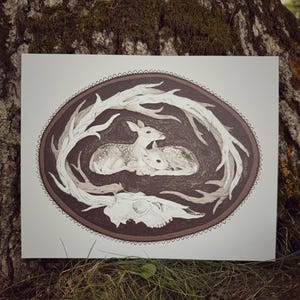 May include: An oval-shaped art print featuring two spotted fawns nestled together, surrounded by a wreath of branches and a skull at the bottom. The artwork is primarily in shades of brown and white, with a detailed, nature-inspired design.