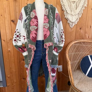 May include: A long-sleeved open-front cardigan featuring a woven pattern of red apples and green leaves on a cream background. The garment has a dark green border, frayed fringe trim along the edges, and two front pockets.