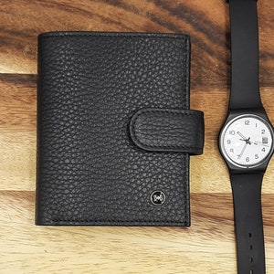 Puede incluir: Una cartera de cuero negro con cierre de solapa y un logotipo plateado. Un reloj de pulsera negro con esfera blanca y agujas plateadas est&aacute; al lado de la cartera.