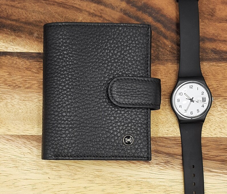 Puede incluir: Una cartera de cuero negro con cierre de solapa y un logotipo plateado. Un reloj de pulsera negro con esfera blanca y agujas plateadas est&aacute; al lado de la cartera.