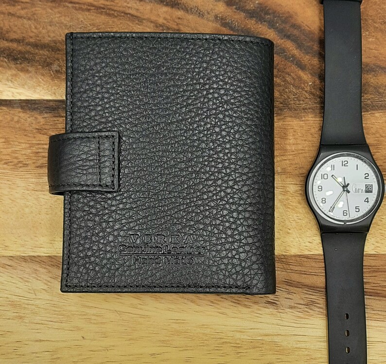 Puede incluir: Una cartera de cuero negro con cierre de solapa y un logotipo plateado en relieve que dice "V.D.R.A. Genuine Leather Hand Made".