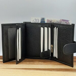 Puede incluir: Cartera de cuero negro de tres pliegues con m&uacute;ltiples ranuras para tarjetas, un compartimento con cremallera y un cierre a presi&oacute;n. La cartera est&aacute; abierta y se puede ver un billete de veinte libras en el interior.