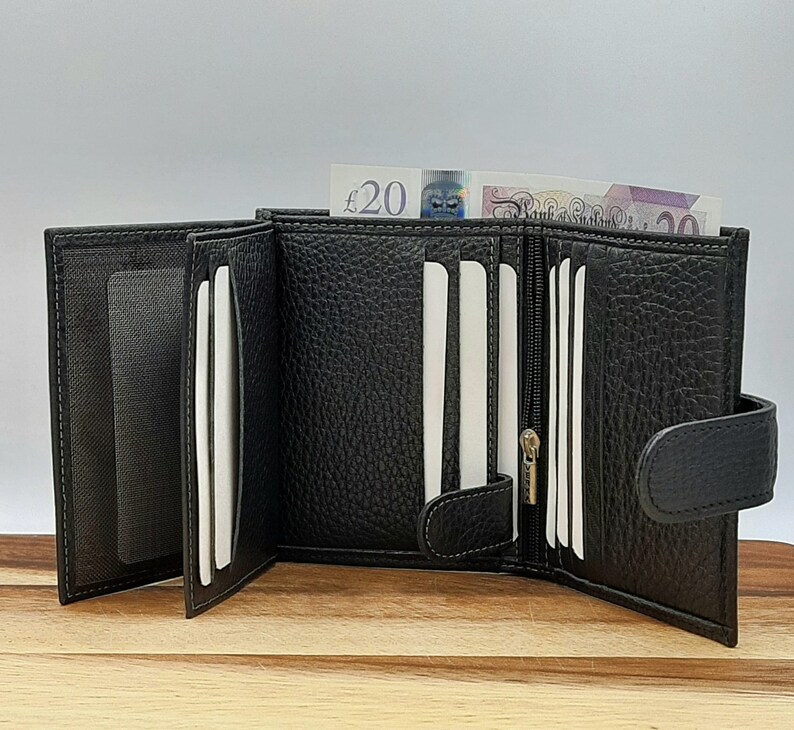 Puede incluir: Cartera de cuero negro de tres pliegues con m&uacute;ltiples ranuras para tarjetas, un compartimento con cremallera y un cierre a presi&oacute;n. La cartera est&aacute; abierta y se puede ver un billete de veinte libras en el interior.