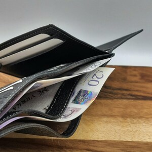 Puede incluir: Una cartera de cuero negro con detalles de costura gris est&aacute; abierta y llena de billetes de veinte libras.